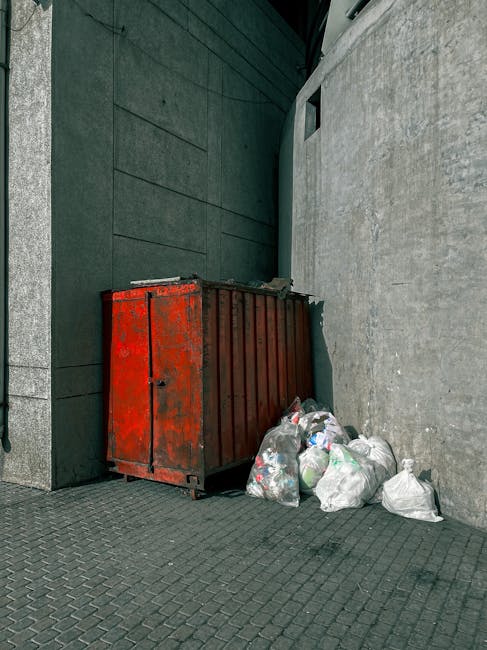 A row of three large wheeled rubbish bins positioned on a paved residential sidewalk beside a brick house wall. The bins are made of black plastic with textured surfaces and closed lids, featuring yel